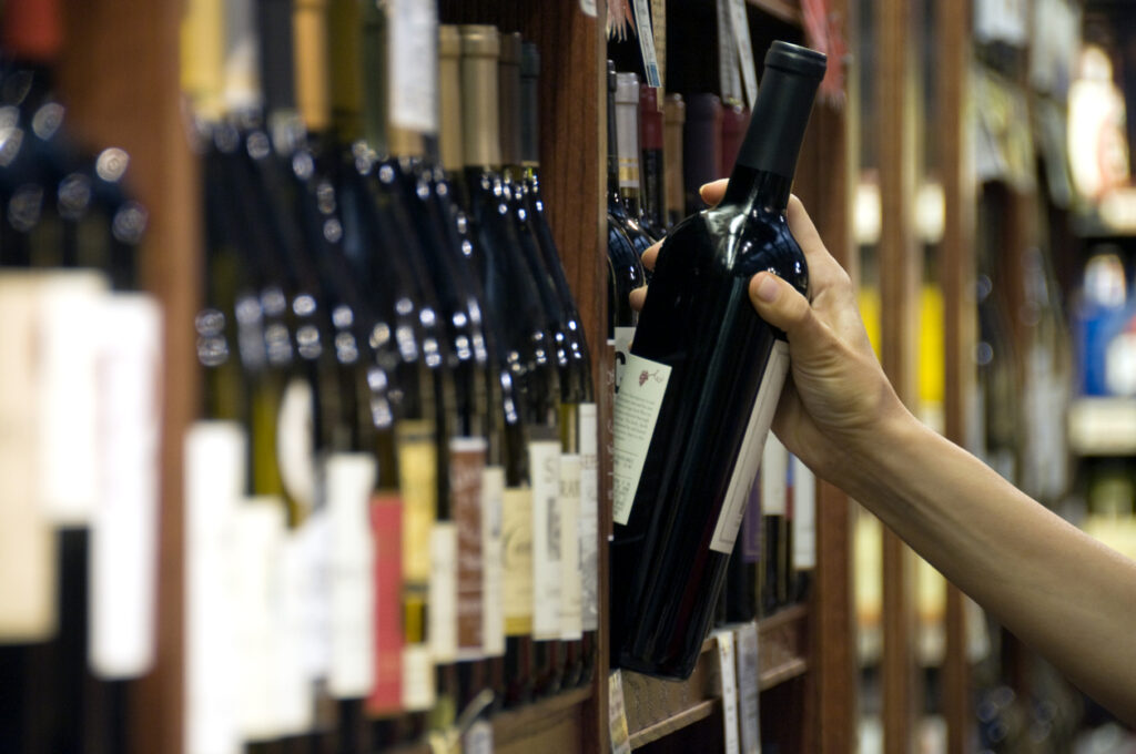 A woman's hand reaches out to select a bottle of red wine from the shelf of a wine shop
