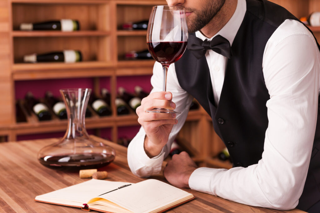 Cropped image of confident male sommelier examining wine while smelling it and leaning at the wooden table with wine shelf in the background