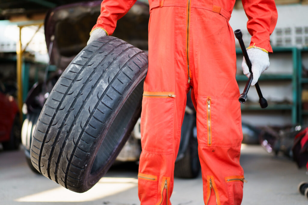 Portrait of professional Asian male vehicle technician or repairman in orange suit holding - carrying a damaged vehicle tyre to be a fixed, a repair man fixing and recap a broken tyre in garage.